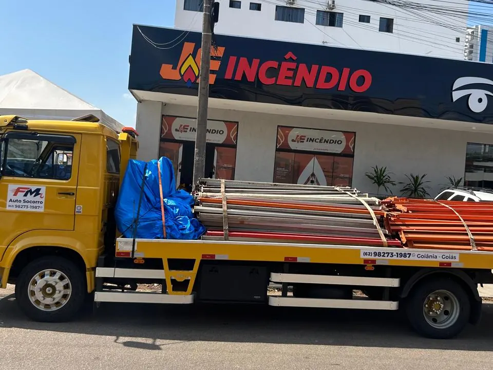Vista lateral do caminhão plataforma da FM Auto Socorro com estruturas metálicas amarradas e lona azul, em frente à loja JE Incêndio