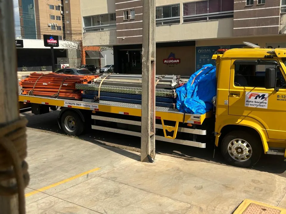 Caminhão plataforma amarelo da FM Auto Socorro carregado com estruturas metálicas laranja e cinza e volumes cobertos por lona azul, parado na rua